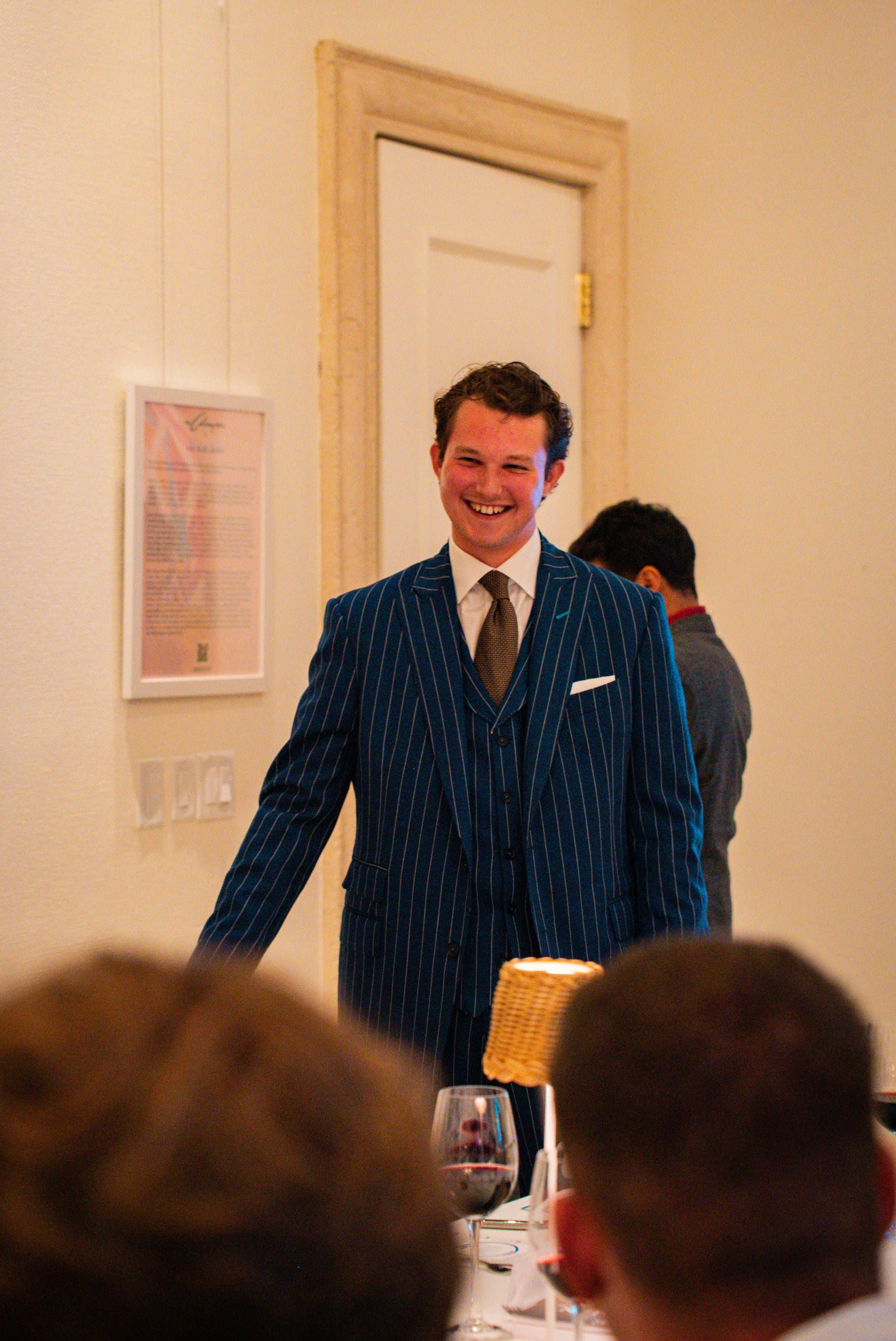 Man in a blue pinstripe suit standing in a room with people and decor.
