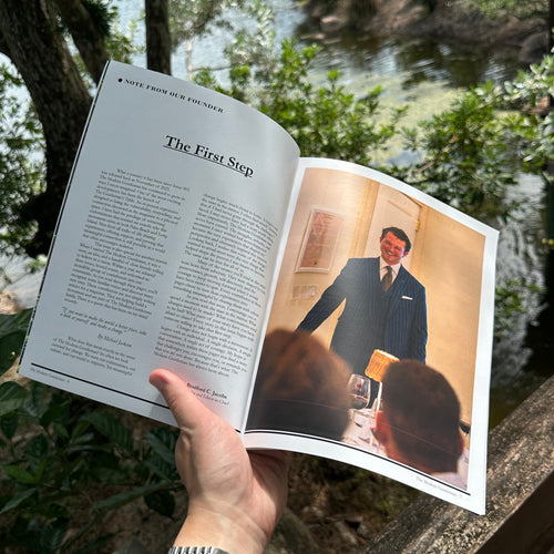 A hand holds an open printed booklet or magazine outdoors near a river, with trees and water visible in the background. The booklet pages are visible, including a photo of a man in a suit on the right page.