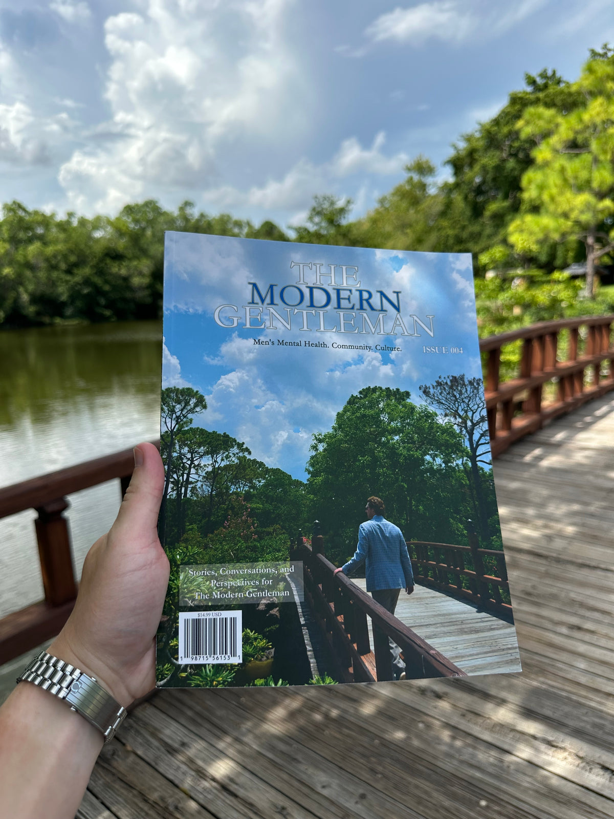 A hand holds a copy of The Modern Gentleman magazine against an outdoor background of a wooden boardwalk and a lake.